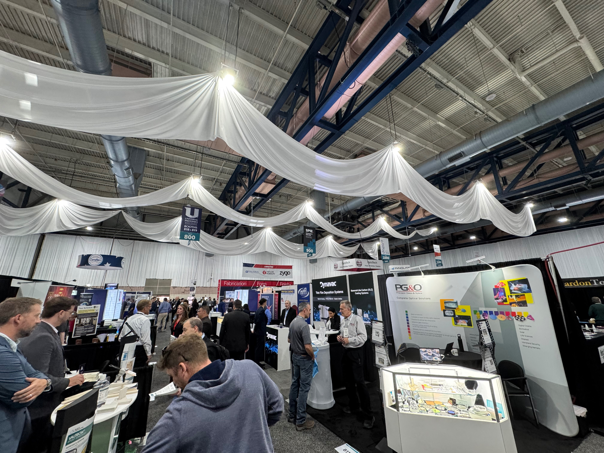 SPIE Optifab 2025 show floor in Rochester with PG&O Precision Glass & Optics, Dynavac, and Universal Photonics booths under draped ceiling